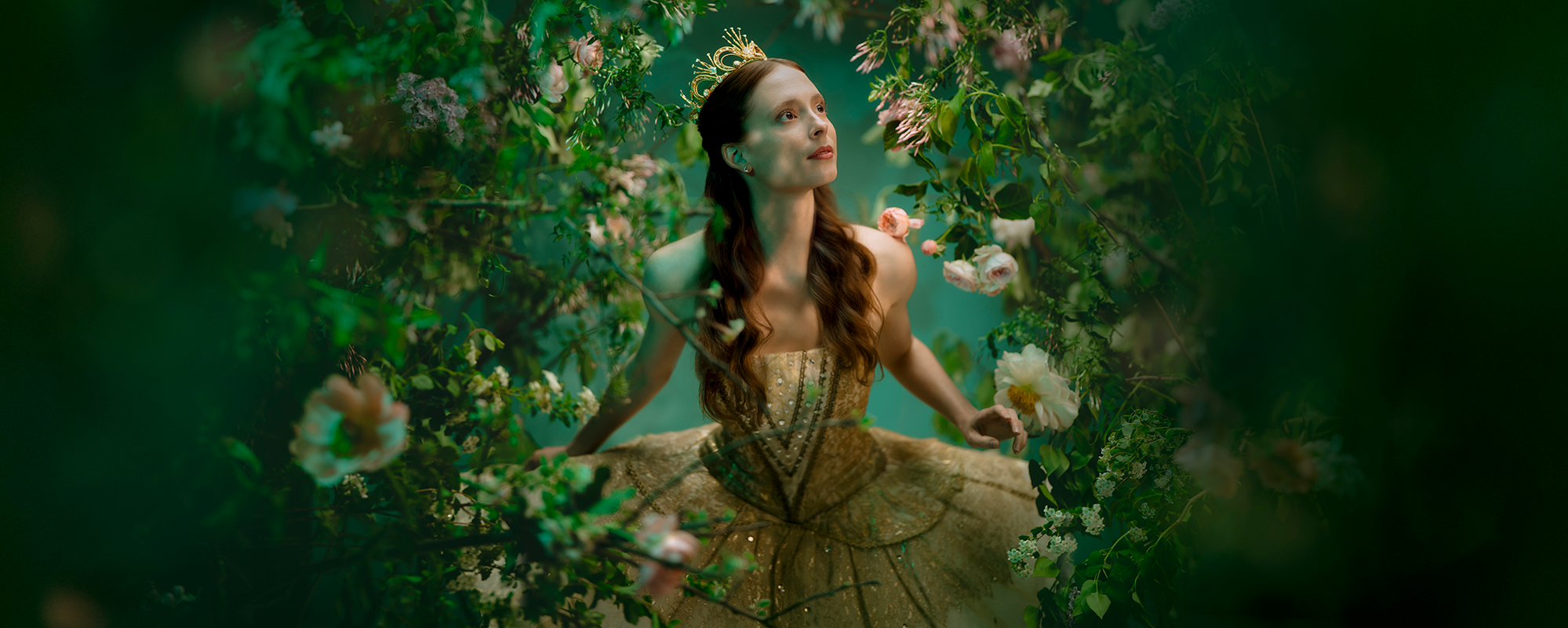 A ballerina dressed as The Sleeping Beauty's Princess Aurora in a gold tutu and crown stands among lush green foliage and flowers, gazing upward as if discovering the enchanted forest around her.