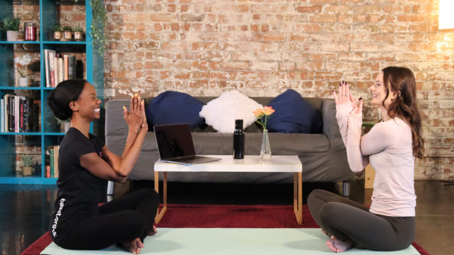A dance tutor and a demonstrator are sitting on a yoga mat in a living room. They are smiling while doing an arms stretch.