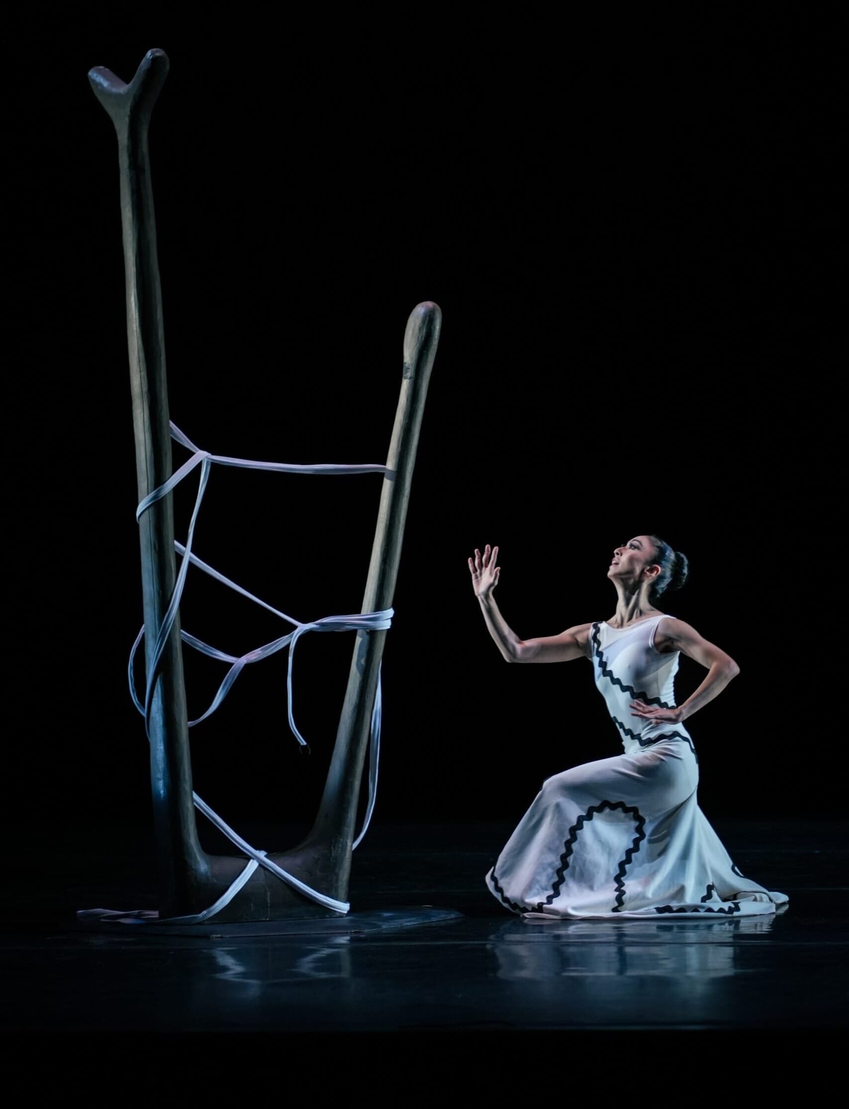 A modern dancer kneels in a white dress with black trim, reaching toward a large wooden structure wrapped in ribbons, set against a black stage.