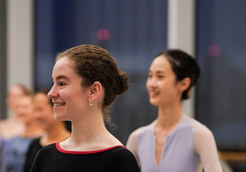 A dancer wearing a black long sleeve leotard stands with her arms rounded by her waist, she is smiling and looking towards the teacher. In the background other dancers stand in the same position. They are in a ballet studio, with ballet barres in the background.