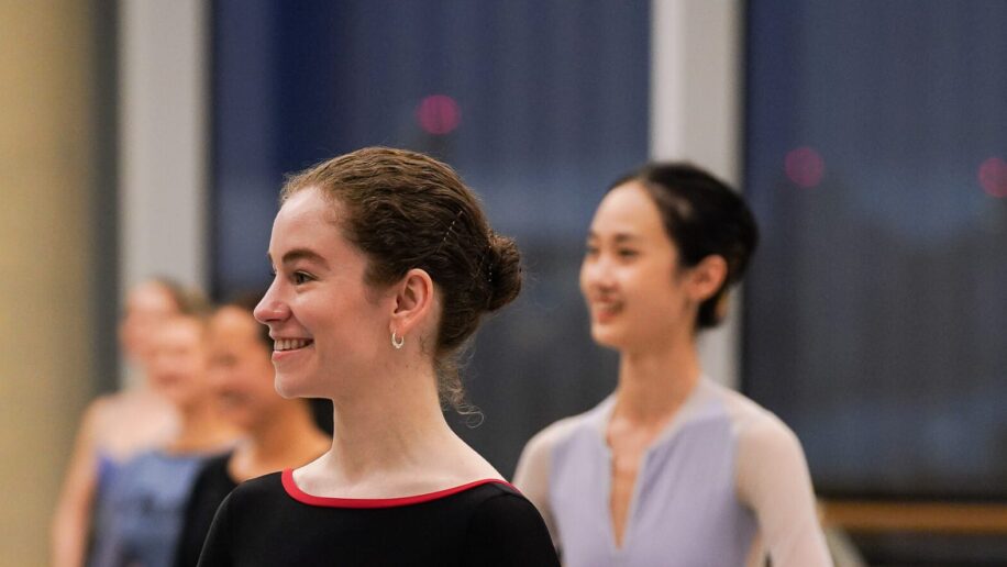 A dancer wearing a black long sleeve leotard stands with her arms rounded by her waist, she is smiling and looking towards the teacher. In the background other dancers stand in the same position. They are in a ballet studio, with ballet barres in the background.