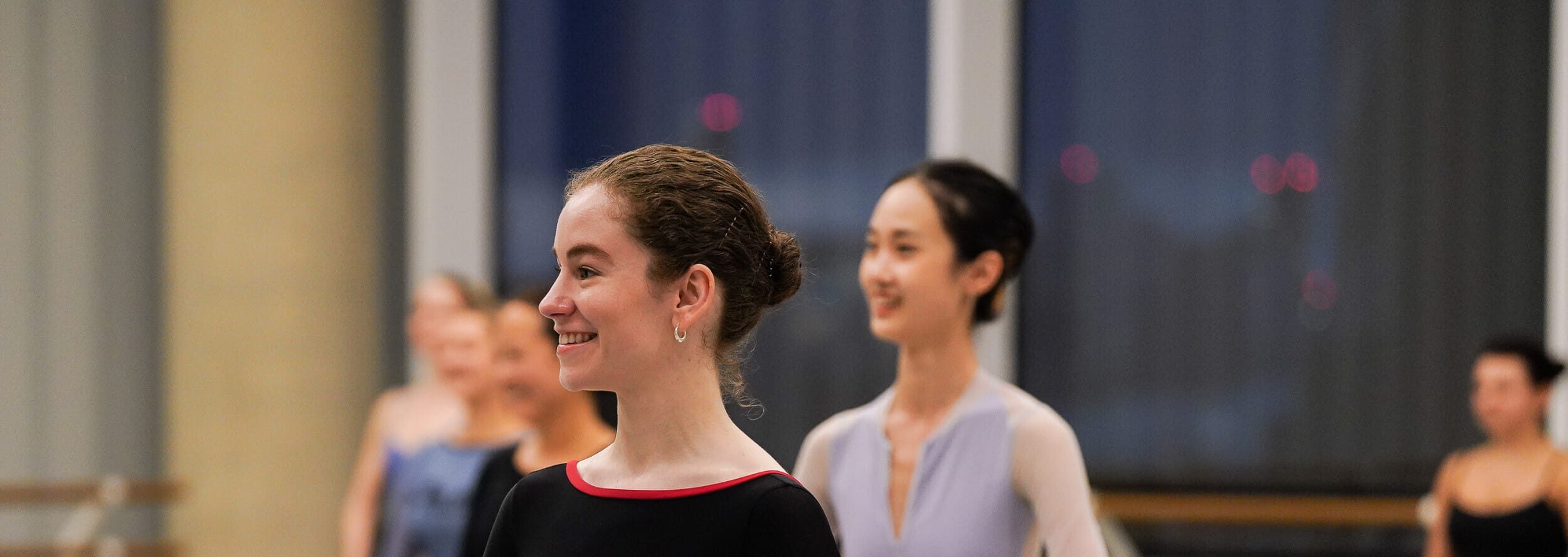 A dancer wearing a black long sleeve leotard stands with her arms rounded by her waist, she is smiling and looking towards the teacher. In the background other dancers stand in the same position. They are in a ballet studio, with ballet barres in the background.