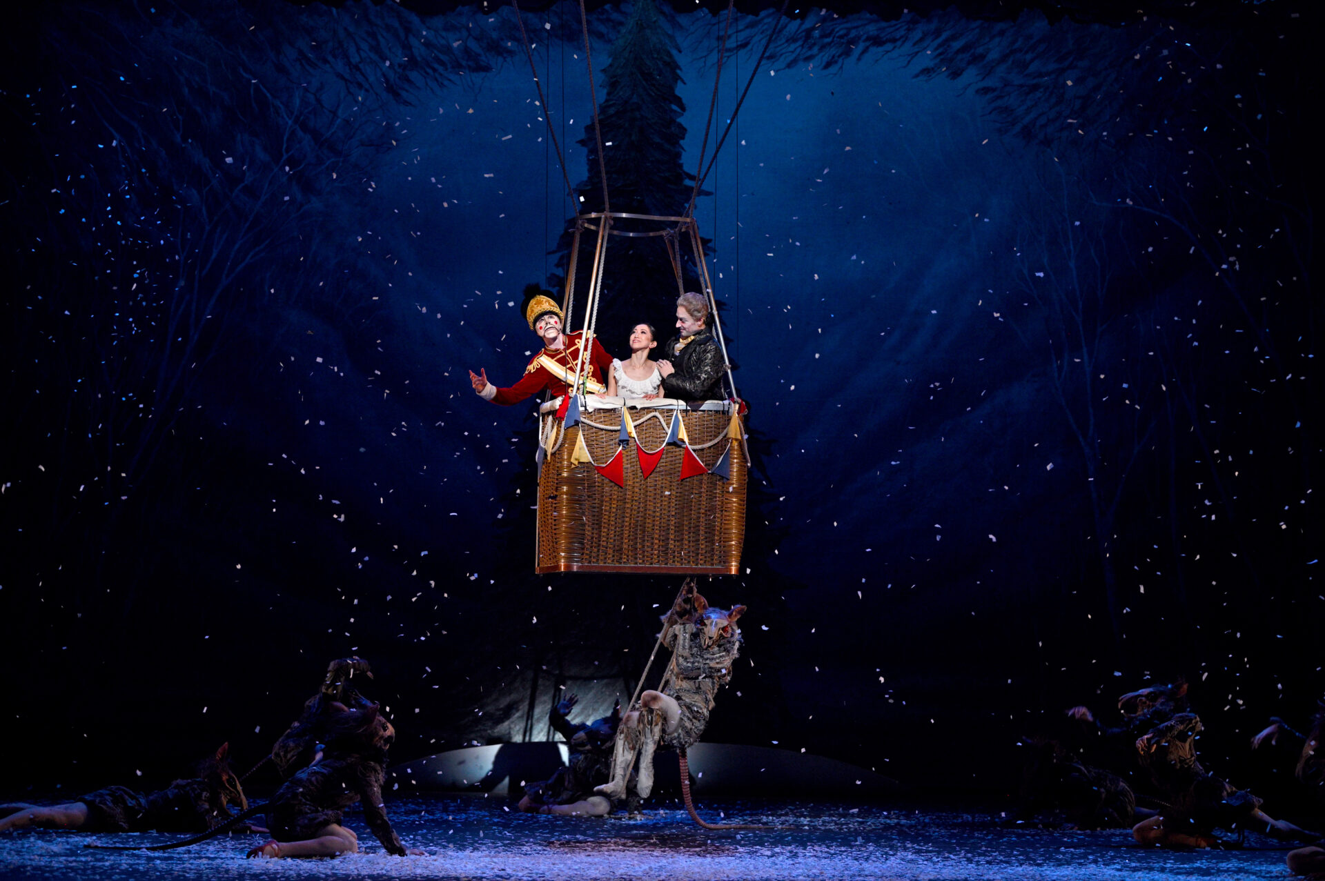 A colourful stage scene shows three performers in a hot air balloon basket, suspended in the air. One performer is dressed as a Nutcracker soldier in red and gold, while the other two, a woman in a white gown, portraying Clara, and an older man in dark attire, portraying Drosselmeyer, look on with joy. The background is a dark, snowy forest, with dancers dressed as mice below.