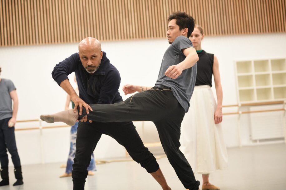 Choreographer assisting a male dancer in a rehearsal studio, with another dancer observing in the background. The studio has modern lighting, wooden panel accents, and ballet barres along the walls.