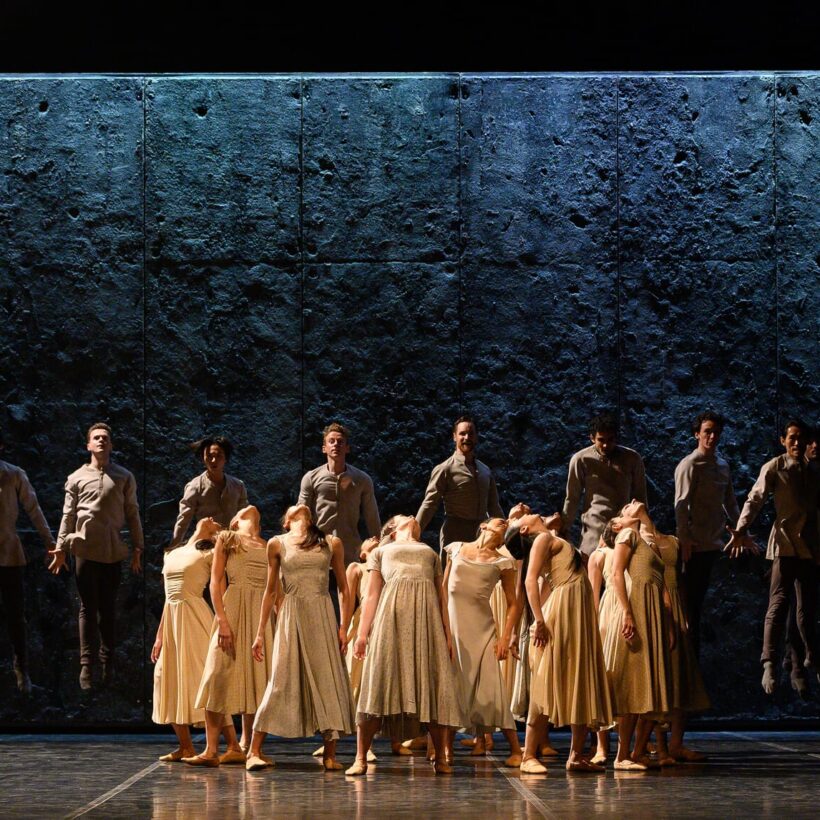 A group of ballet dancers from Akram Khan’s Giselle is divided into two distinct formations on stage. In the foreground, female dancers in cream-colored dresses gather tightly together, their heads thrown back in unison. Behind them, a line of male dancers dressed in grey coats hovers midair against a dark, textured backdrop.