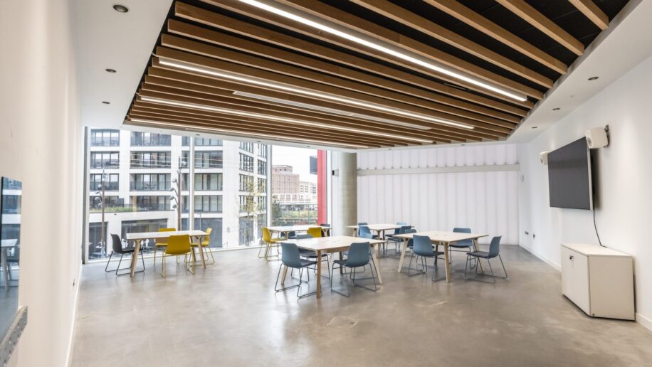 Modern meeting room with large windows, natural light, and a view of city buildings. The room features a stylish ceiling with wooden beams, multiple tables and chairs in a minimalistic setup, and a large wall-mounted screen.