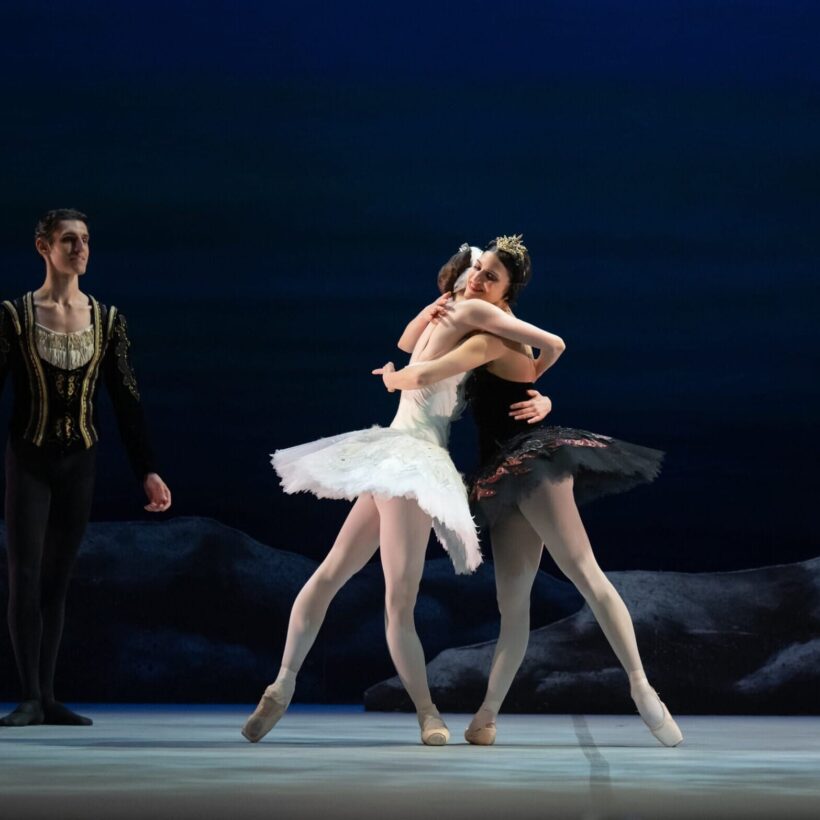 Ballet performance scene featuring three dancers: a ballerina in a white tutu as Odette, a ballerina in a black tutu as Odile embracing Odette, and a male dancer in a princely costume as Prince Siegfried. The background depicts a serene, dark blue setting with rocky elements, evoking a lakeside atmosphere.