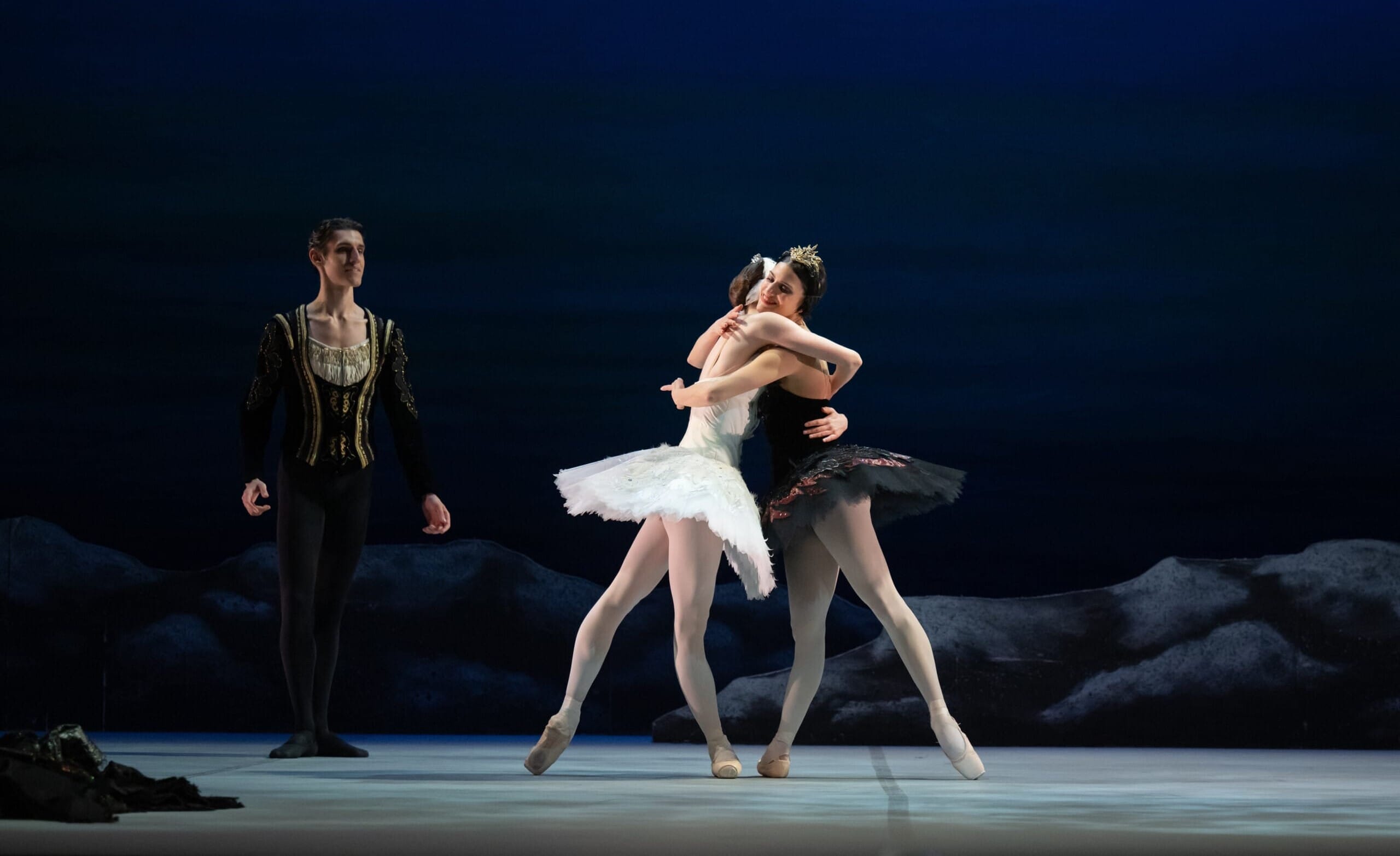 Ballet performance scene featuring three dancers: a ballerina in a white tutu as Odette, a ballerina in a black tutu as Odile embracing Odette, and a male dancer in a princely costume as Prince Siegfried. The background depicts a serene, dark blue setting with rocky elements, evoking a lakeside atmosphere.