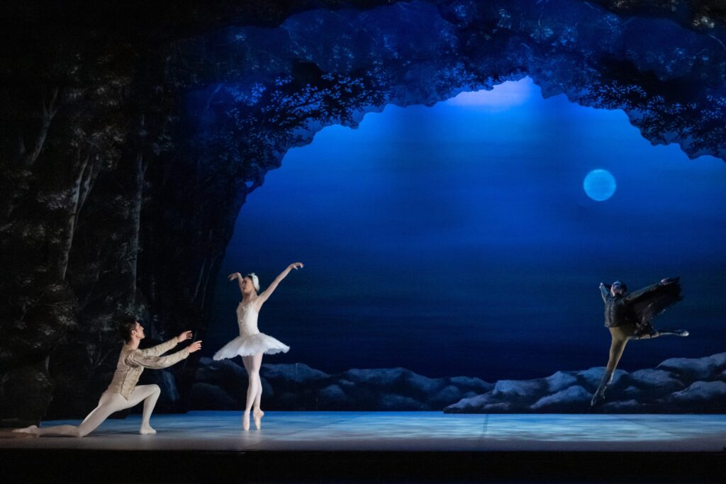 A dramatic ballet scene set under a glowing moonlit sky. On the left, a male dancer as Prince Siegfried kneels with arms outstretched towards a ballerina in a white tutu as Odette, who stands gracefully in pointe. On the right, a dancer as Rothbart leaps dynamically with outspread arms and a dark, wing-like costume. The stage is framed by rocky scenery and an arched silhouette of trees.
