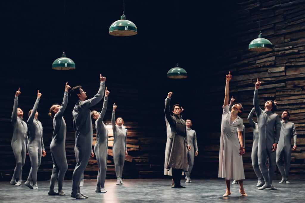 A group of dancers is shown pointing upwards towards the ceiling, four lamps hang above them
