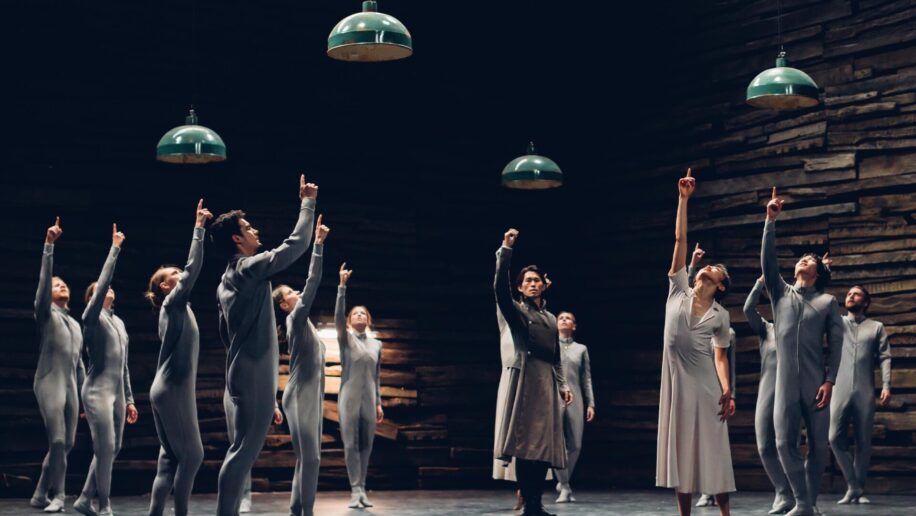 A group of dancers is shown pointing upwards towards the ceiling, four lamps hang above them