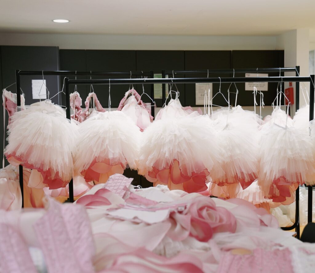 A clothing rack holding multiple delicate ballet tutus from Nutcracker in shades of white and soft pink. In the foreground, additional pieces of pink fabric and costume elements lie on a table, suggesting a costume workshop setting.