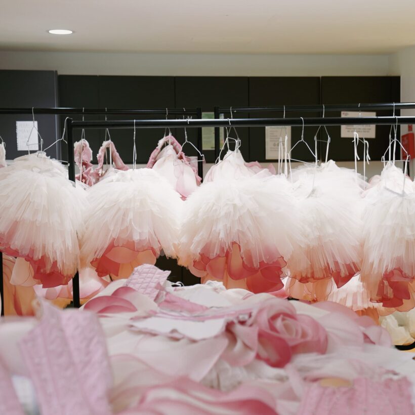 A clothing rack holding multiple delicate ballet tutus from Nutcracker in shades of white and soft pink. In the foreground, additional pieces of pink fabric and costume elements lie on a table, suggesting a costume workshop setting.