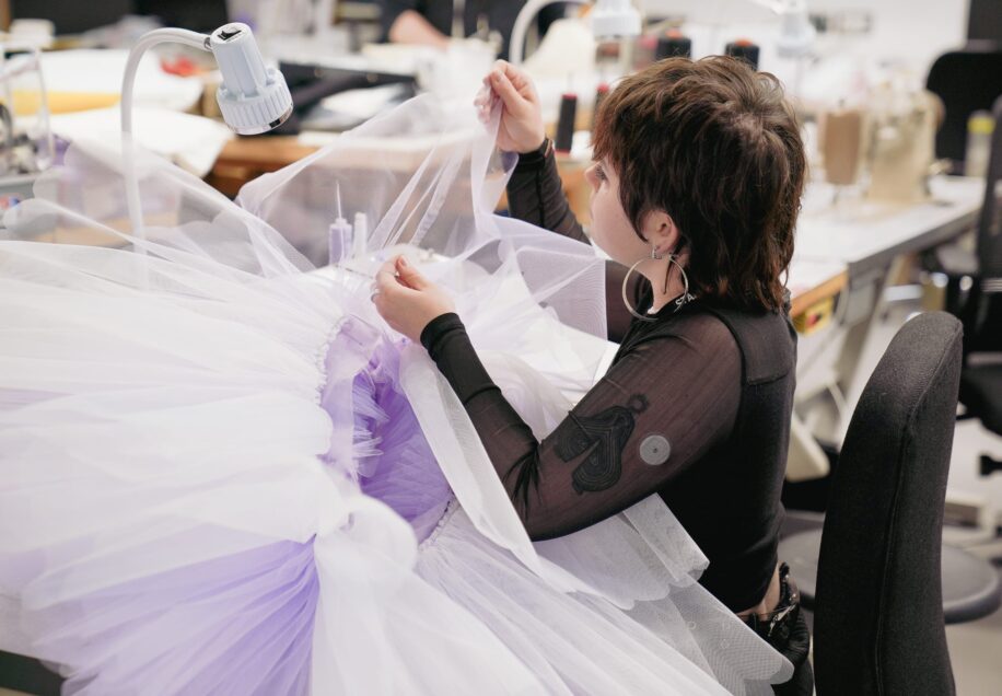 A costume maker carefully sewing layers of tulle onto a ballet tutu in shades of white and lavender for the Sugar Plum Fairy in Nutcracker. The maker, wearing a black mesh top with visible tattoos on their arm, works with focus under a bright desk lamp in a workshop filled with sewing equipment.