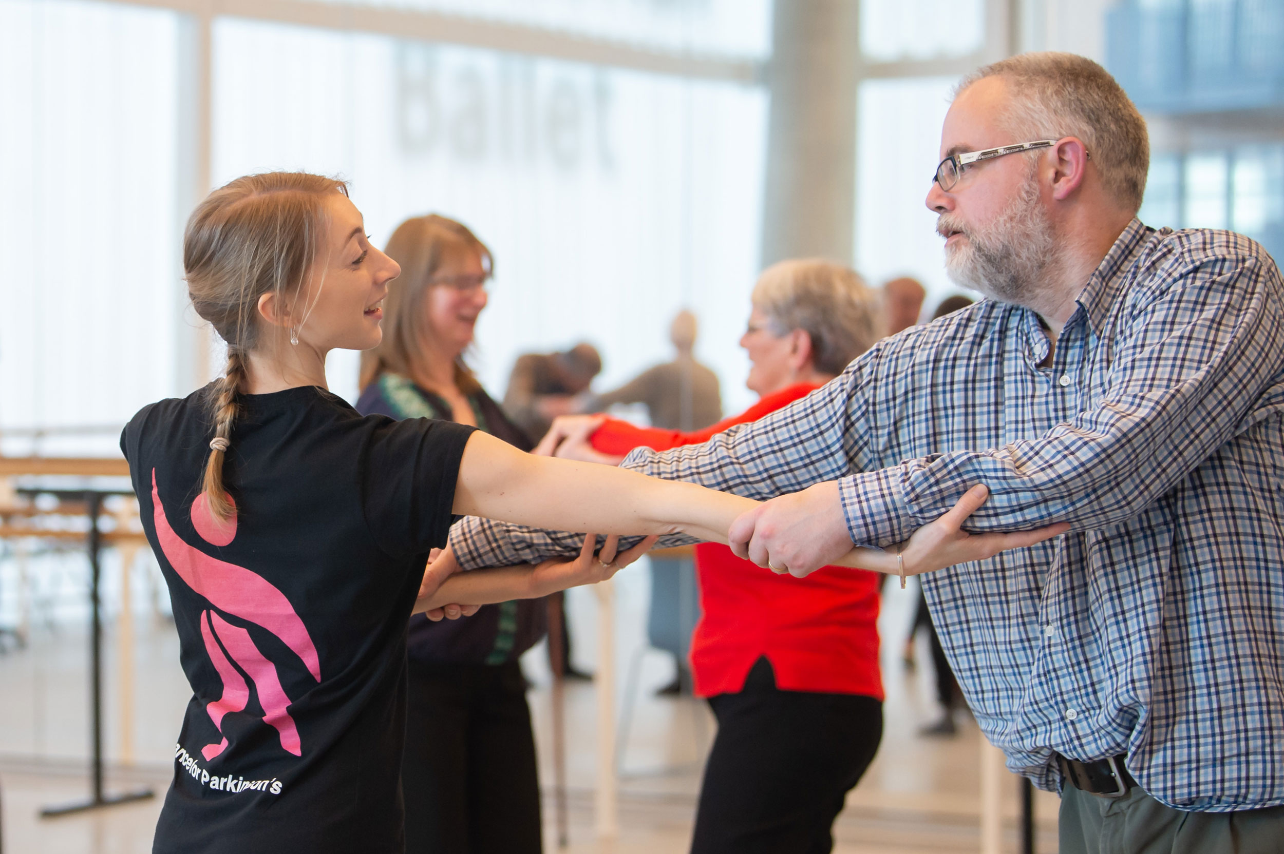 A dance teacher wearing a black t-shirt is dancing with an older man wearing a plaid shirt. They are holding hands and arms in a dance pose. In the background, other people can be seen participating in a dance class in a bright, windowed room.