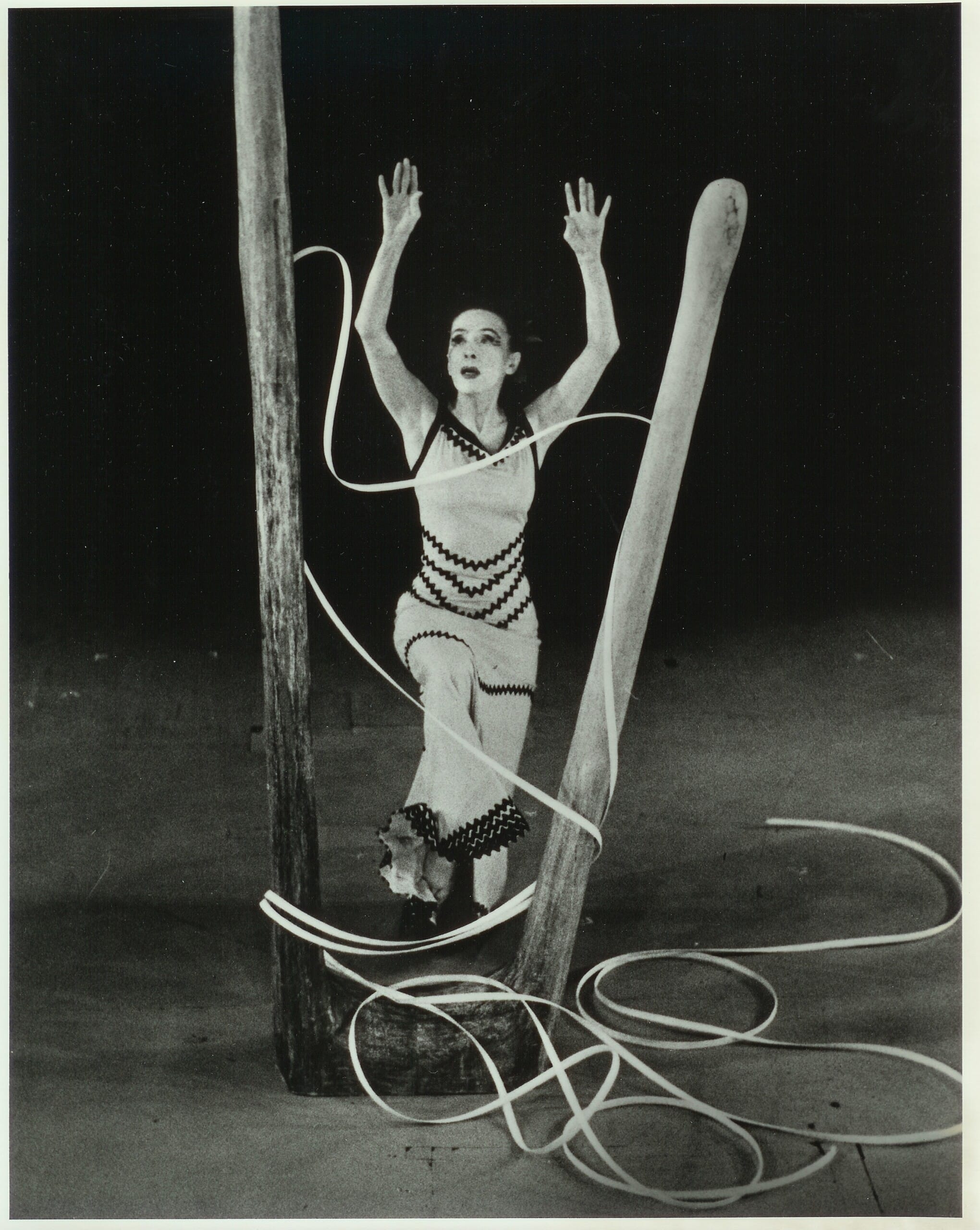 A dancer performs on stage surrounded by large wooden structures and long, coiled ropes. She is dressed in a form-fitting costume with zigzag patterns, one knee raised, and arms extended upward, conveying a sense of motion and intensity against a dark background.
