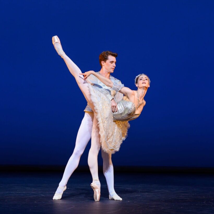 A classical ballet dancer in a sparkling tutu is held in an elegant arabesque by her partner against a deep blue backdrop.