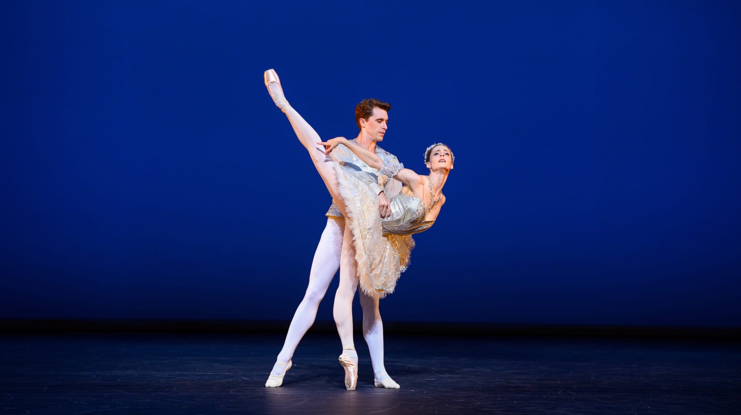 A classical ballet dancer in a sparkling tutu is held in an elegant arabesque by her partner against a deep blue backdrop.