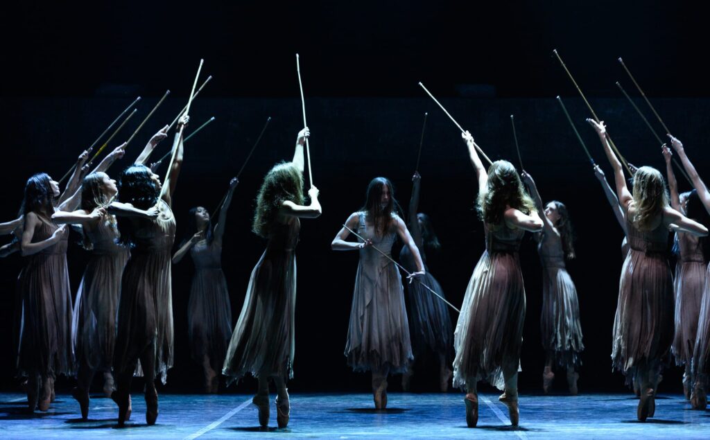 Ballet dancers, dressed in flowing, tattered beige costumes, perform on stage in an intense, ritualistic scene from Akram Khan’s Giselle. They hold long wooden sticks aloft, reaching towards the sky. The dimly lit stage creates a haunting and atmospheric effect. The central figure, with long hair obscuring her face, stands out, moving with commanding strength.