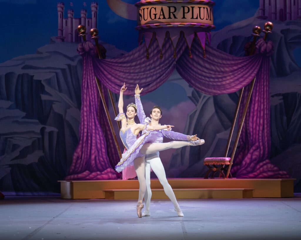 Two ballet dancers perform a graceful duet on stage in a scene from Nutcracker. The female dancer, dressed in a lavender tutu and tiara, is en pointe with one leg extended behind her, supported by her partner in white tights and a matching lavender jacket.