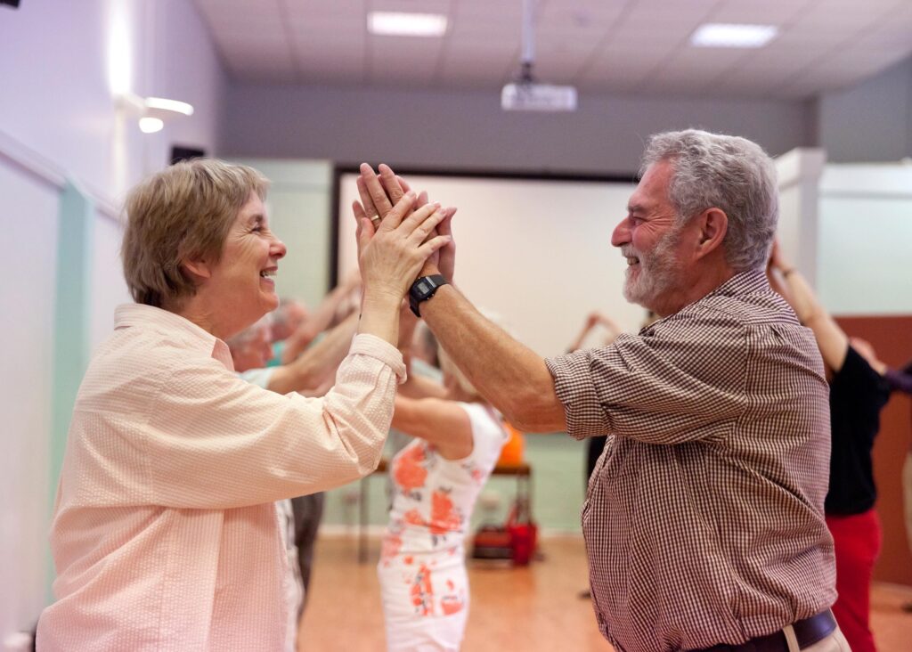 The image shows a joyful moment in a dance class, where a man and woman are smiling at each other while participating in a partnered exercise. They are facing each other, lightly touching their hands together as part of the dance activity. The scene takes place in a well-lit room, with other participants visible in the background. The atmosphere is one of warmth, connection, and enjoyment, reflecting the social and physical benefits of dance classes for individuals with Parkinson's.