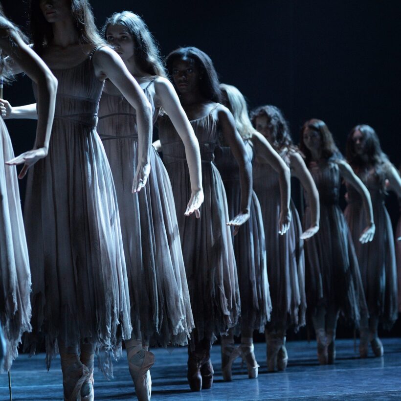 English National Ballet’s dancers performs as Wilis from Akram Khan's Giselle. The dancers stand in a line, dressed in tattered dresses, and pose on pointe with their arms extended in a menacing way. The stage is dimly lit, creating a haunting and dramatic atmosphere.