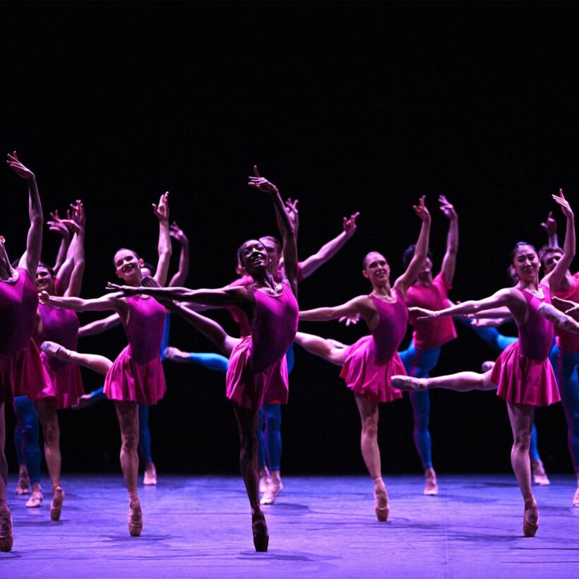 A group of ballet dancers performing in unison in Playlist (EP), part of our 2024/2025 season. The dancers are in a dynamic pose, extending their legs and arms gracefully, with one leg lifted high behind them. The stage is dimly lit, emphasizing their bright pink costumes against a dark background, creating a striking visual contrast.