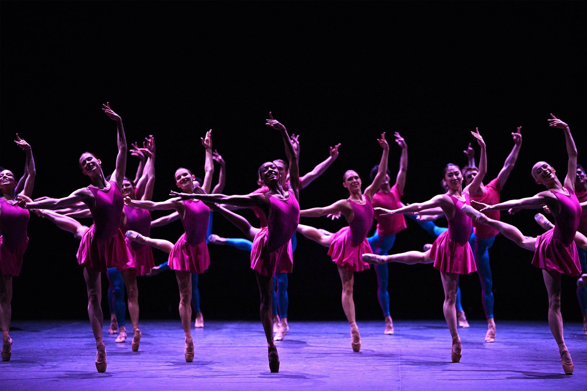 A group of ballet dancers performing in unison in Playlist (EP), part of our 2024/2025 season. The dancers are in a dynamic pose, extending their legs and arms gracefully, with one leg lifted high behind them. The stage is dimly lit, emphasizing their bright pink costumes against a dark background, creating a striking visual contrast.