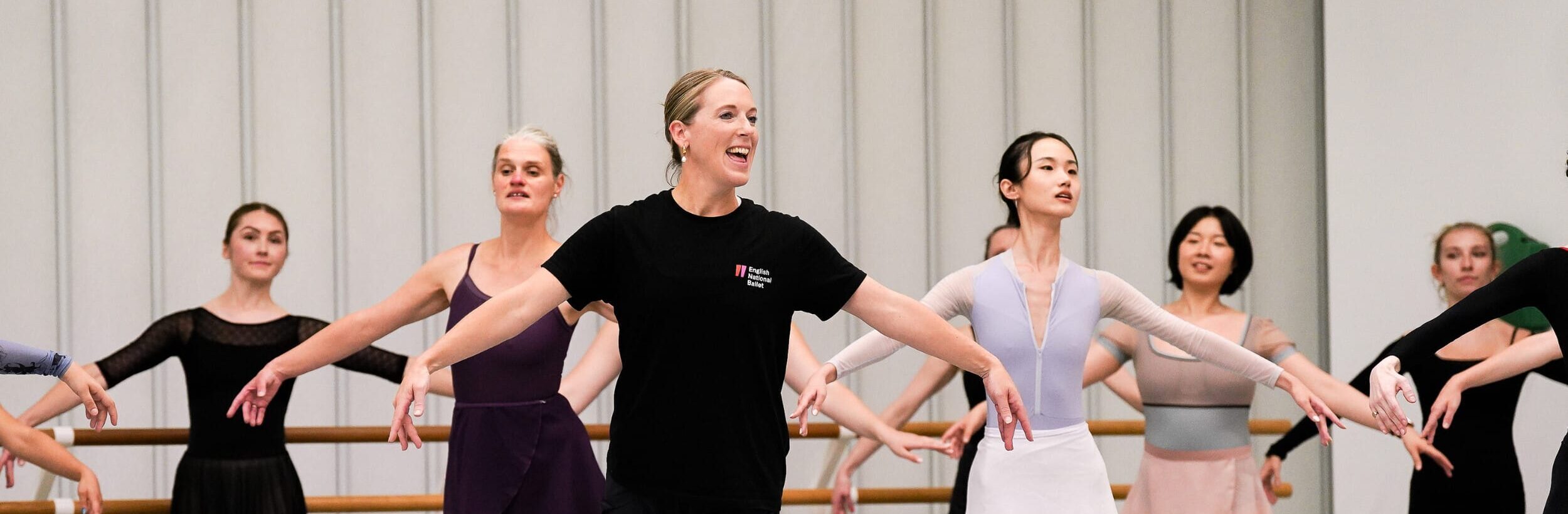A teacher wears all black and stands at the front of a group with her arms outstretched to the side and one leg slightly behind. There are several other dancers who stand behind her, copying her movement.
