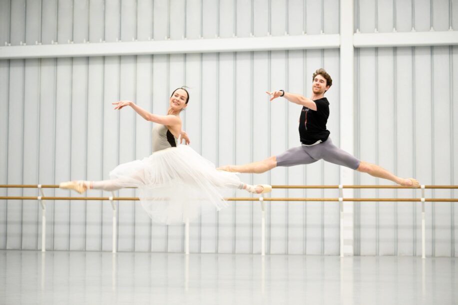 Two ballet dancers are captured mid-air in a rehearsal studio. The female dancer on the left is wearing a long, flowing white tutu and beige leotard, with her legs extended gracefully in a split leap. She has a serene expression, gazing slightly to her right. The male dancer on the right, dressed in a black T-shirt and gray tights, mirrors her movement with a powerful split leap.