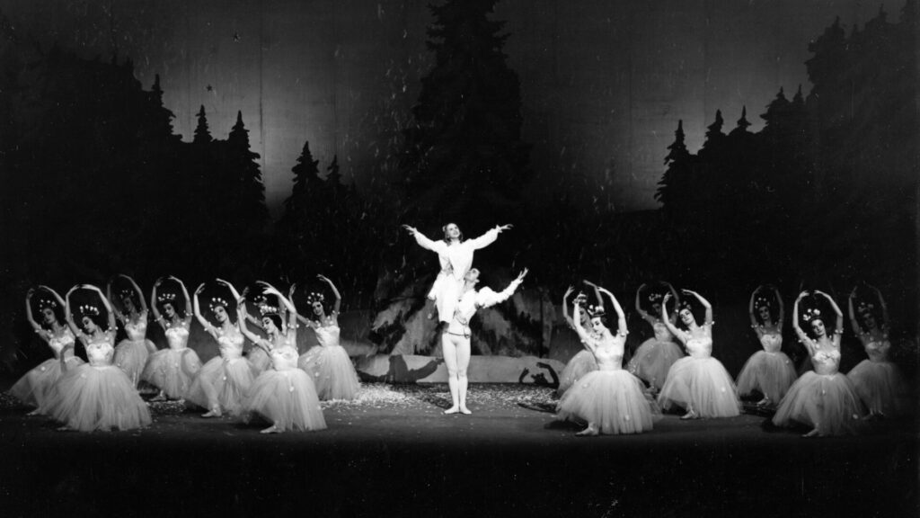 This black-and-white photograph depicts a Nutcracker performance from English National Ballet set in a wintery forest scene. A male dancer lifts a female dancer in the centre, surrounded by a group of ballerinas in tutus and floral headpieces, posed with their arms gracefully arched.