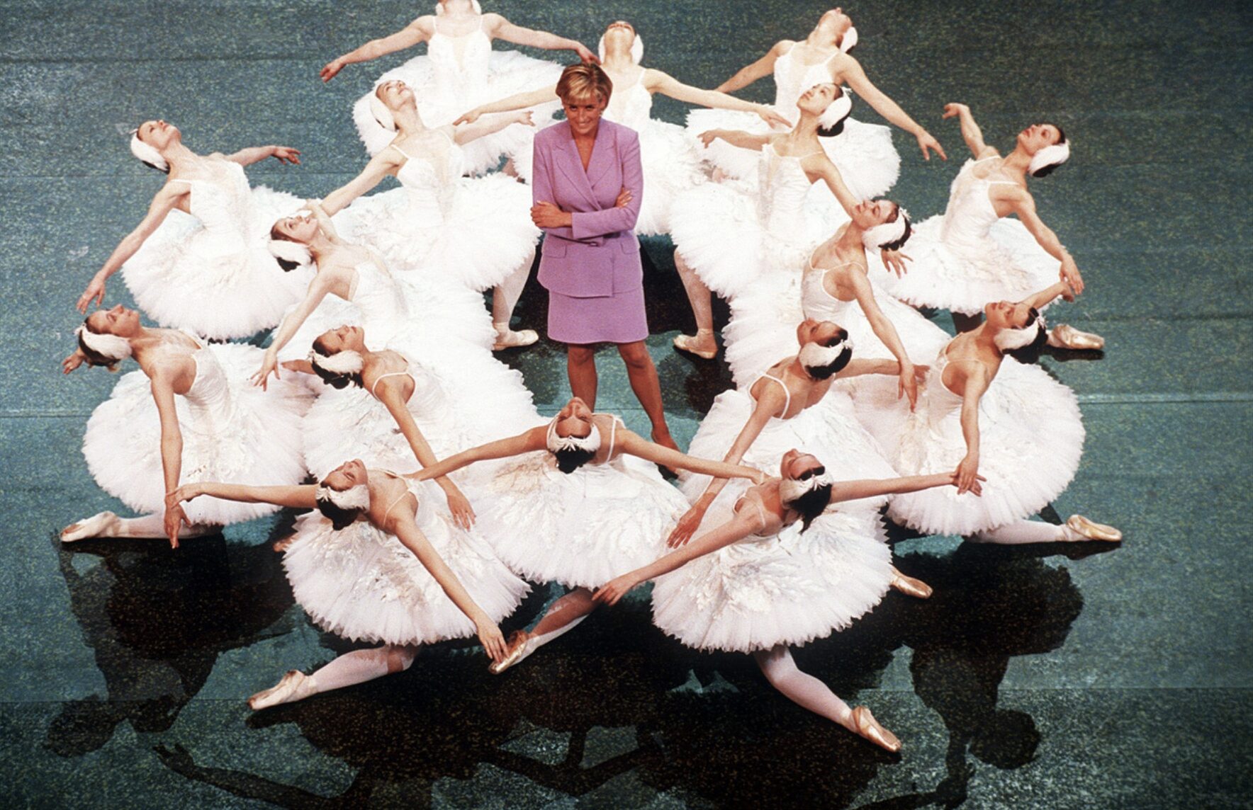 Diana, Princess of Wales is shown wearing a pink dress. 16 white swans corps de ballet dancers are sitting around her, wearing their white tutus.
