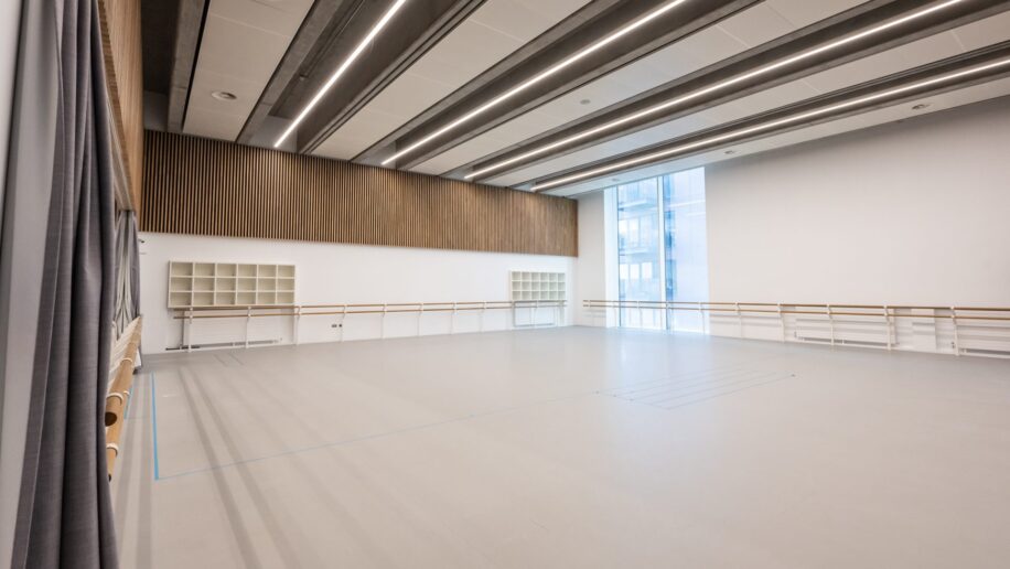 Spacious dance rehearsal studio in London with large windows allowing natural light, wooden barre along the walls, modern lighting, and wooden panel accents on the upper walls.