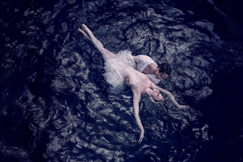 An ethereal scene from Swan Lake: a ballet dancer portraying the White Swan Odette lies gracefully in the arms of her partner, Prince Siegfried, surrounded by dark, rippling water, embodying the beauty and tragedy of the timeless ballet.