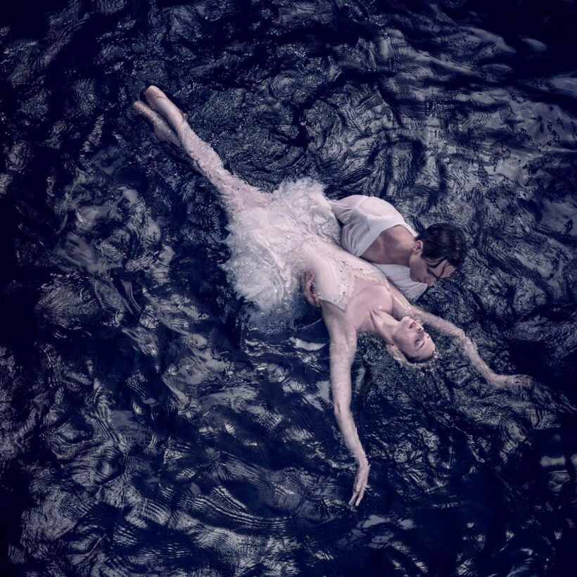 An ethereal scene from Swan Lake: a ballet dancer portraying the White Swan Odette lies gracefully in the arms of her partner, Prince Siegfried, surrounded by dark, rippling water, embodying the beauty and tragedy of the timeless ballet.