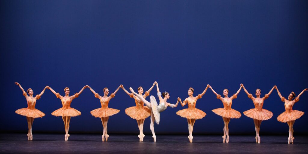 A group of ballet dancers performs on stage in matching orange tutus, with a central dancer in white striking an arabesque pose.