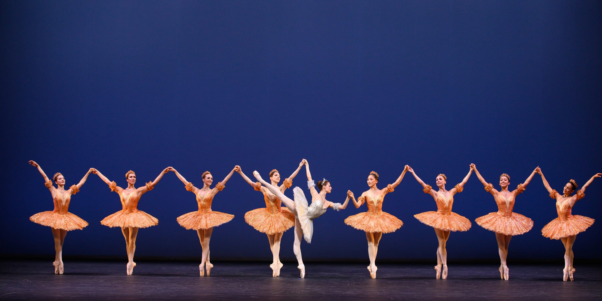 A group of ballet dancers performs on stage in matching orange tutus, with a central dancer in white striking an arabesque pose.
