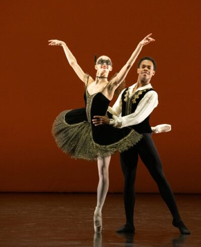 Ballet dancers performing a pas de deux on stage at Emerging Dancer 2025. A female dancer in a black tutu with gold trim executes an attitude en pointe while her male partner in a black and gold vest supports her, set against a warm amber backdrop.