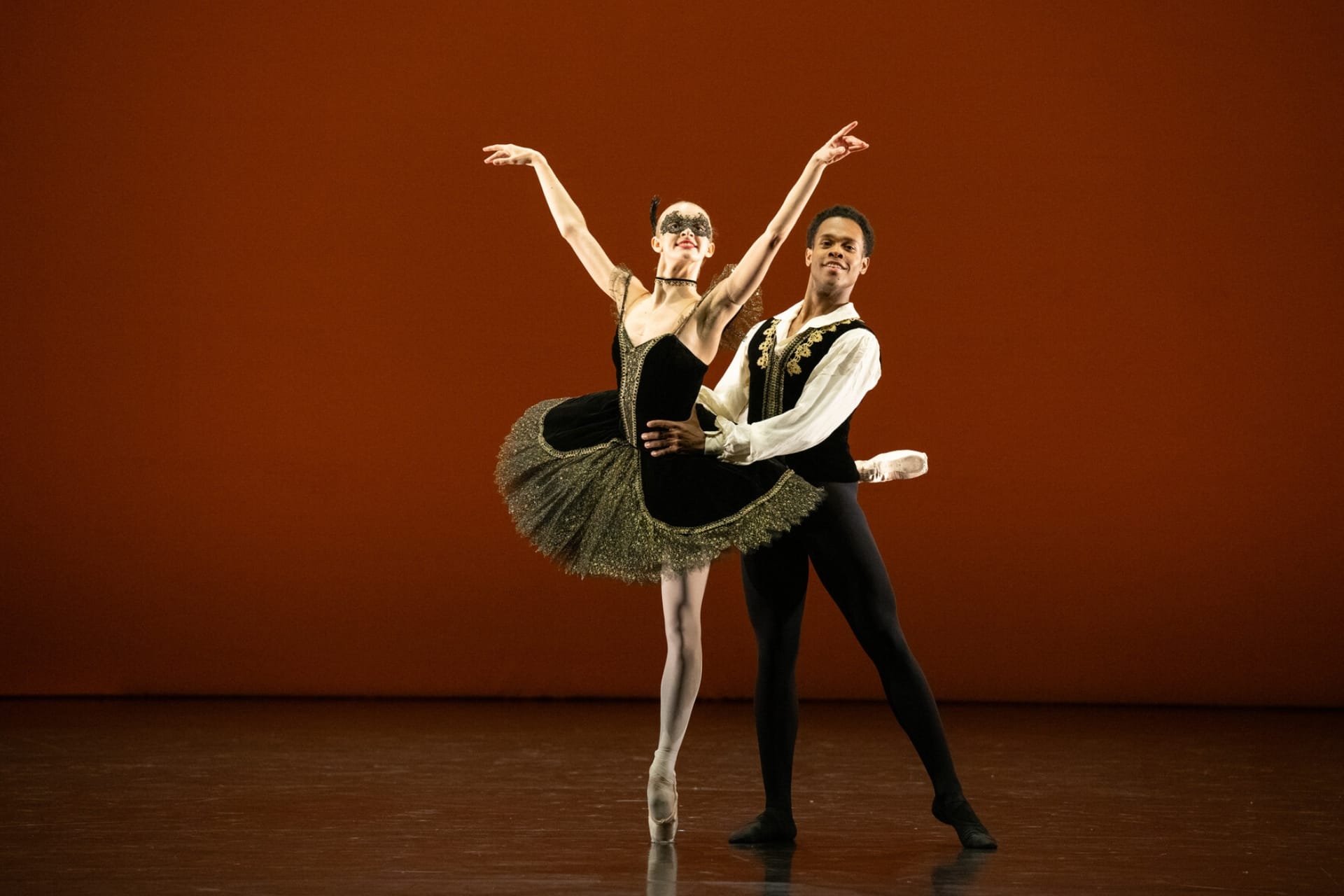 Ballet dancers performing a pas de deux on stage at Emerging Dancer 2025. A female dancer in a black tutu with gold trim executes an attitude en pointe while her male partner in a black and gold vest supports her, set against a warm amber backdrop.