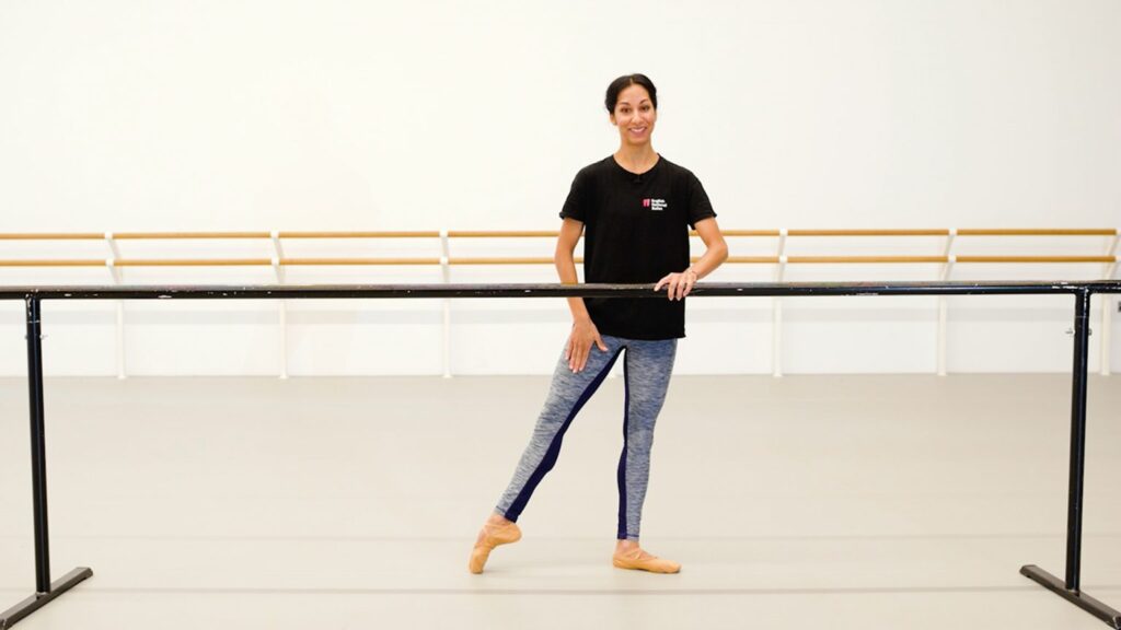 A teacher of dance classes for adults stands in a dance studio, holding onto a ballet barre with one hand while posing with one leg extended to the side. She is smiling and wearing a black t-shirt and blue leggings. The studio has a clean, minimalistic design.
