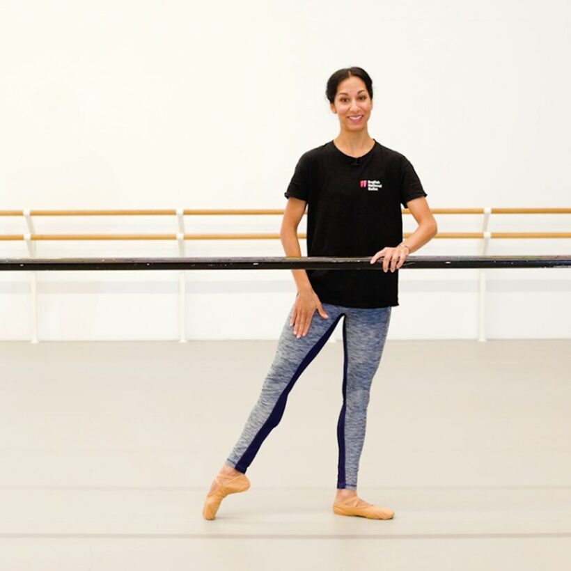 A teacher of dance classes for adults stands in a dance studio, holding onto a ballet barre with one hand while posing with one leg extended to the side. She is smiling and wearing a black t-shirt and blue leggings. The studio has a clean, minimalistic design.