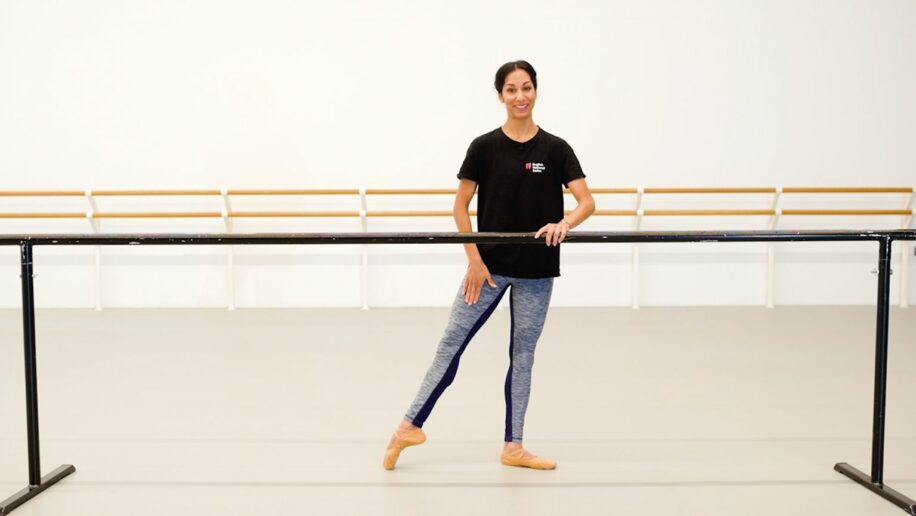A teacher of dance classes for adults stands in a dance studio, holding onto a ballet barre with one hand while posing with one leg extended to the side. She is smiling and wearing a black t-shirt and blue leggings. The studio has a clean, minimalistic design.