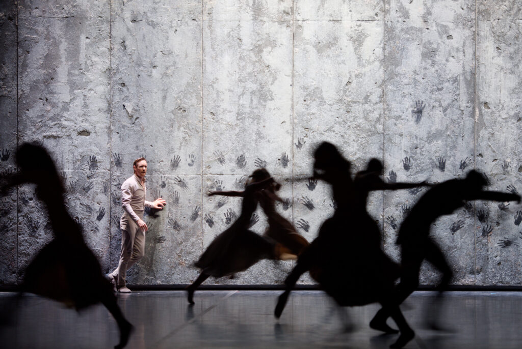A dancer stands with his hand over a wall with other hand prints while the black silhouettes of other dancers are running in the foreground