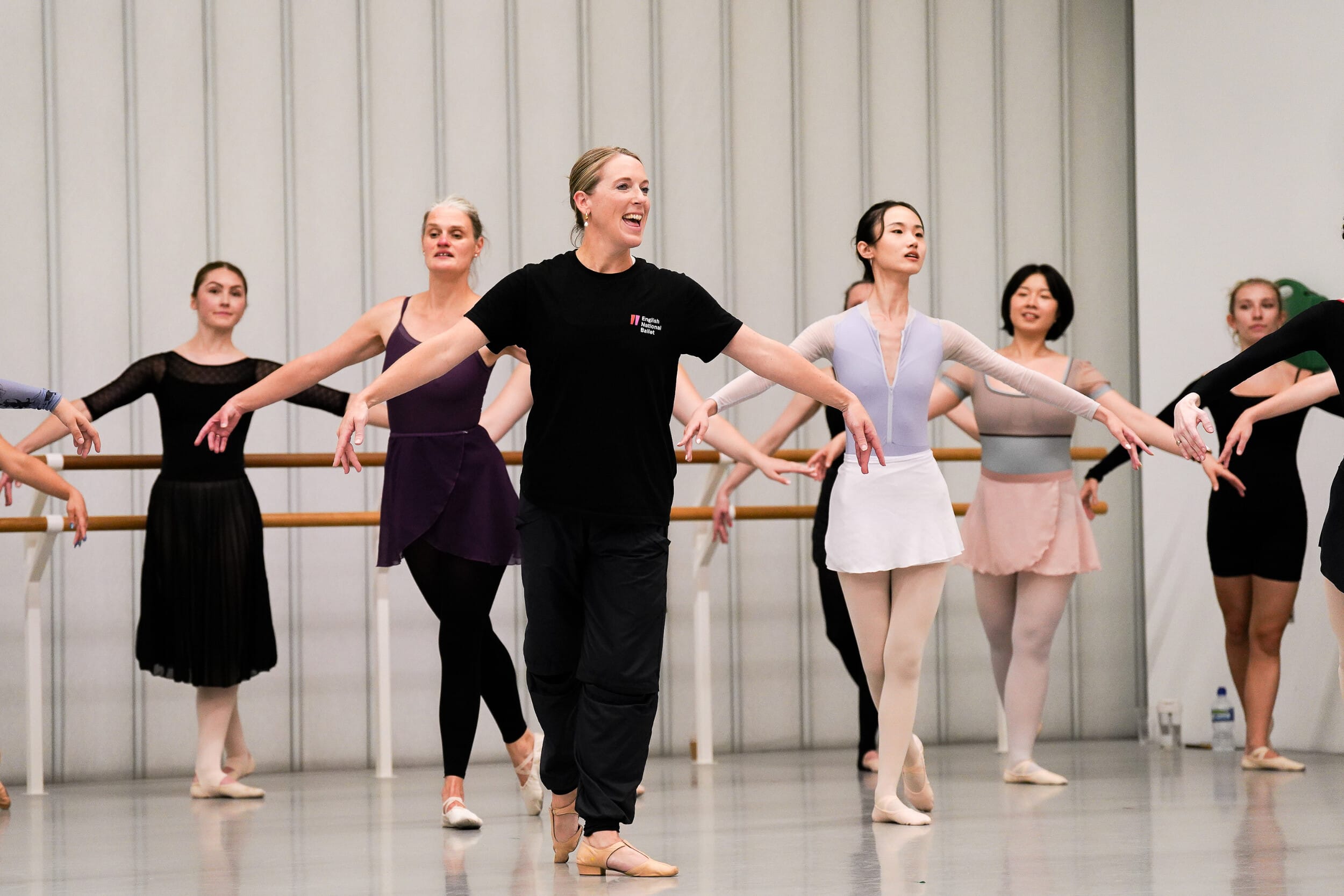 A teacher wears all black and stands at the front of a group with her arms outstretched to the side and one leg slightly behind. There are several other dancers who stand behind her