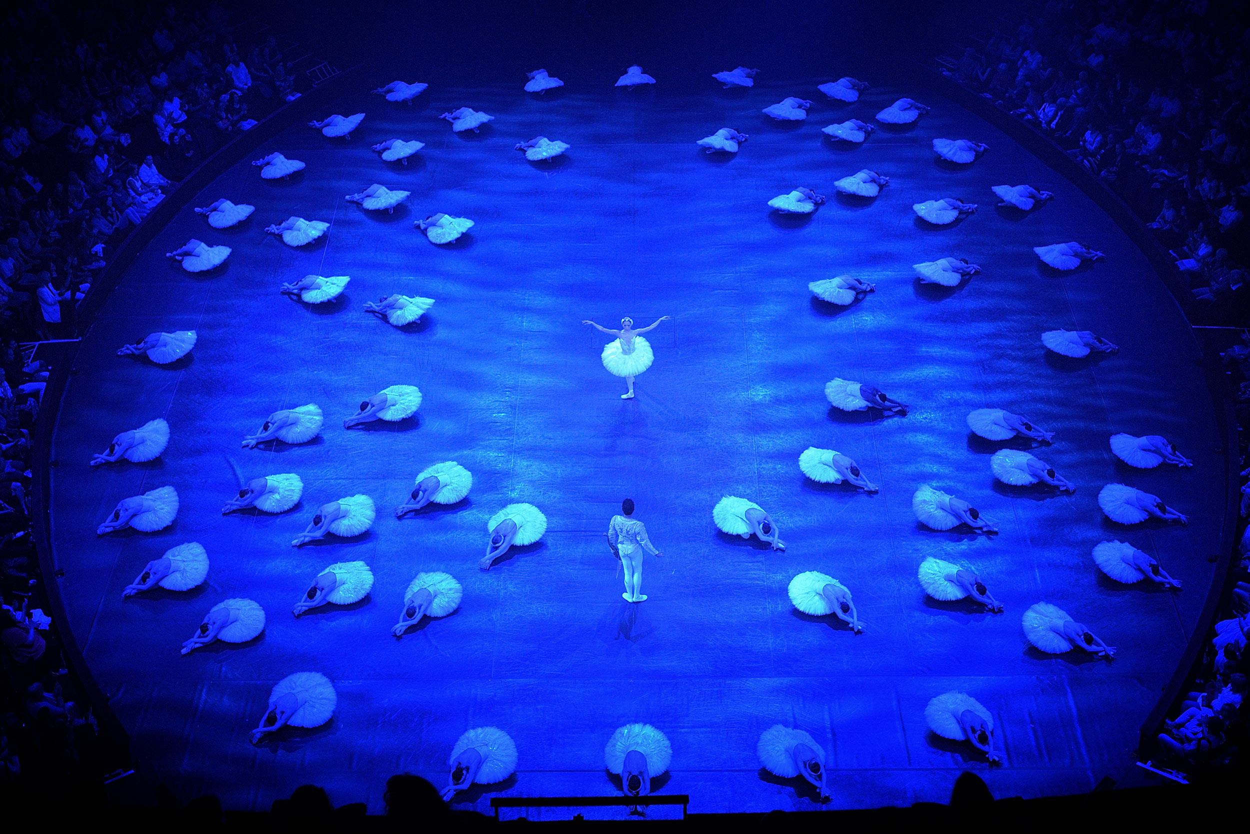 A dancer in the role of Odette from Swan Lake and a dancer in the role of Prince Siegfried are facing each other at the centre of a round stage filled with blue light. Odette is wearing a white tutu and headdress while Siegfried is wearing a white top and white tights. Around them
