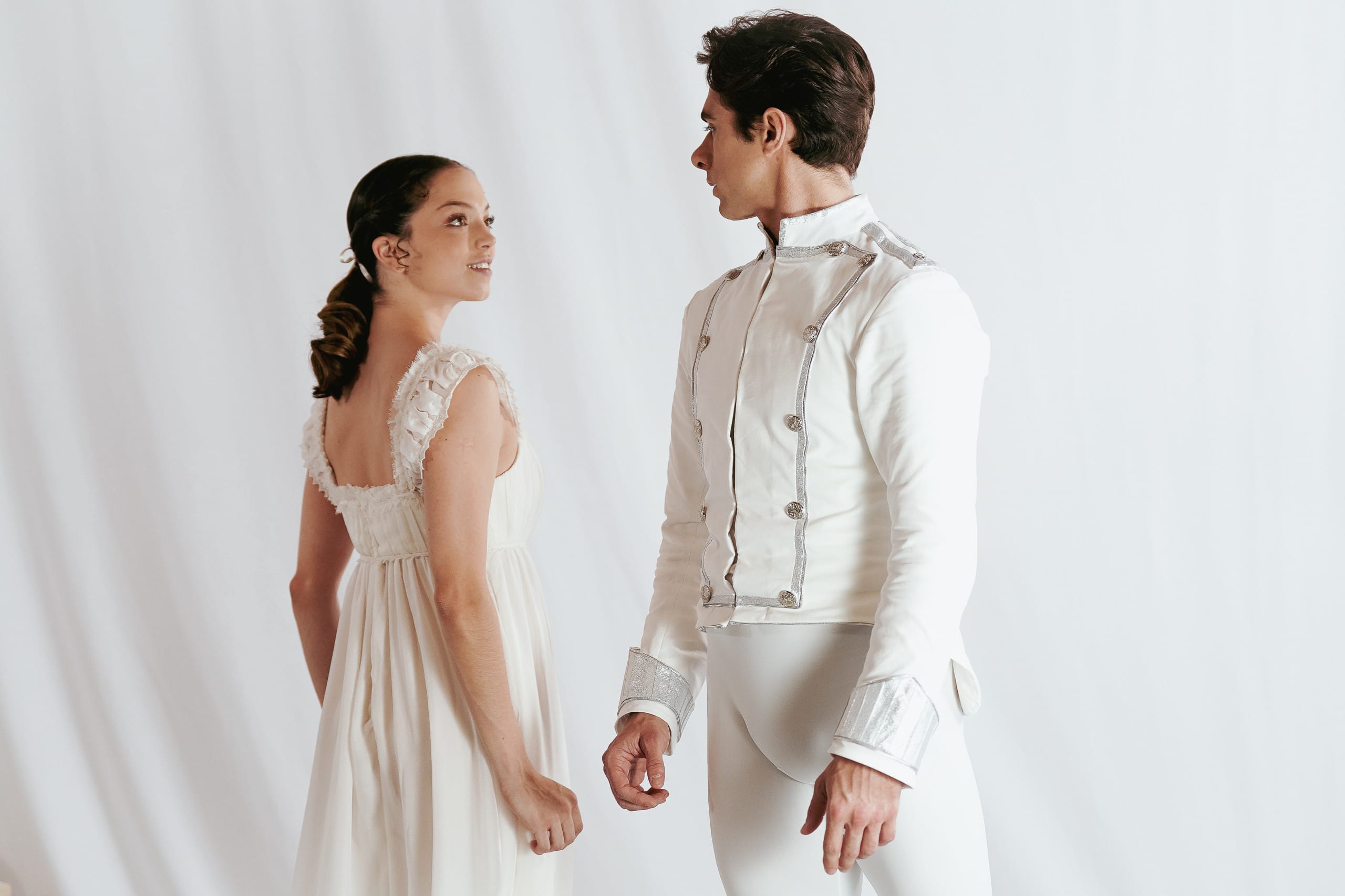 A male and female ballet dancer stand facing each other against a plain white background. The female dancer wears a flowing white dress with delicate ruffled straps