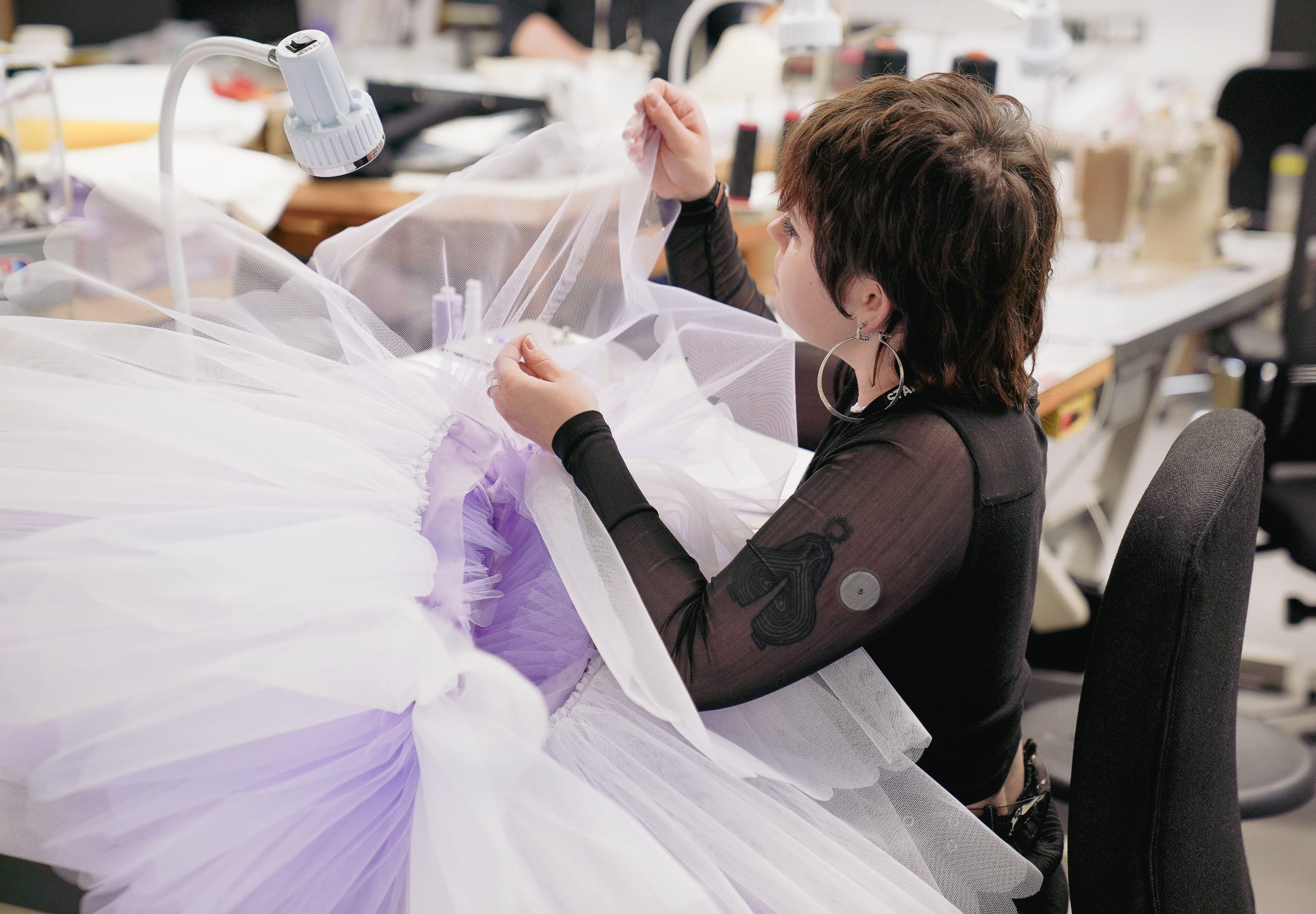 A costume maker carefully sewing layers of tulle onto a ballet tutu in shades of white and lavender for the Sugar Plum Fairy in Nutcracker. The maker