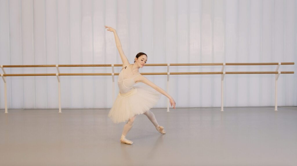 A dancer wearing a white tutu and leotard is shown in a pose from Swan Lake while in the studio.
